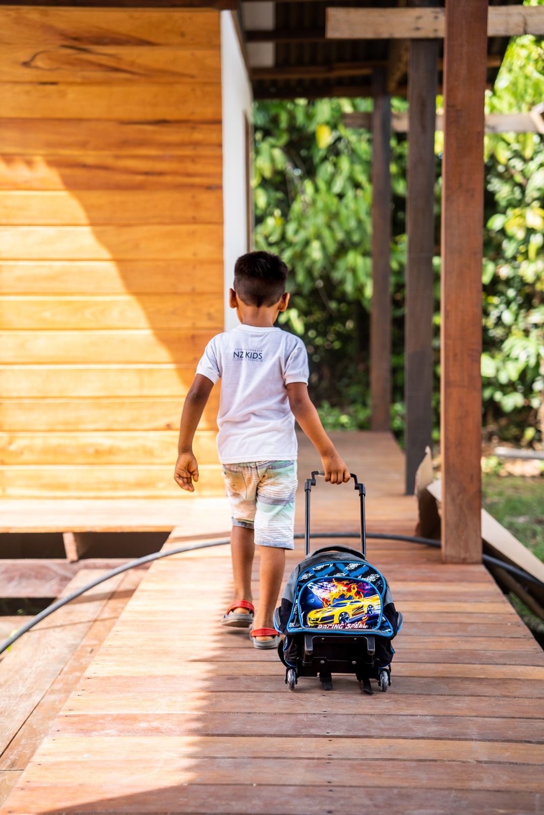 Menino de mochila de rodinhas caminha pela passarela de madeira da escola, cercada de floresta