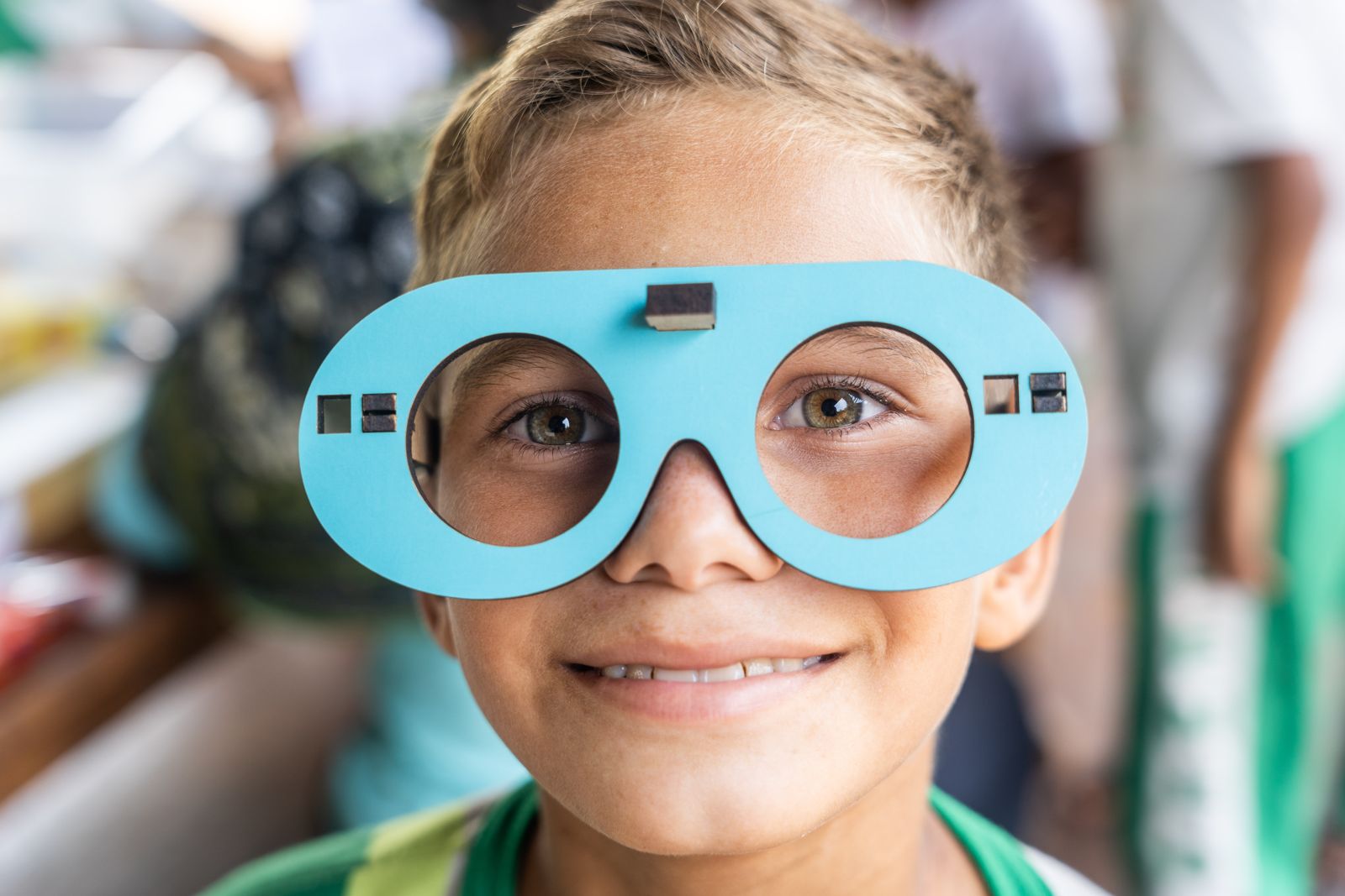 Menino sorridente olha através de uma armação de óculos de madeira azul durante uma atividade