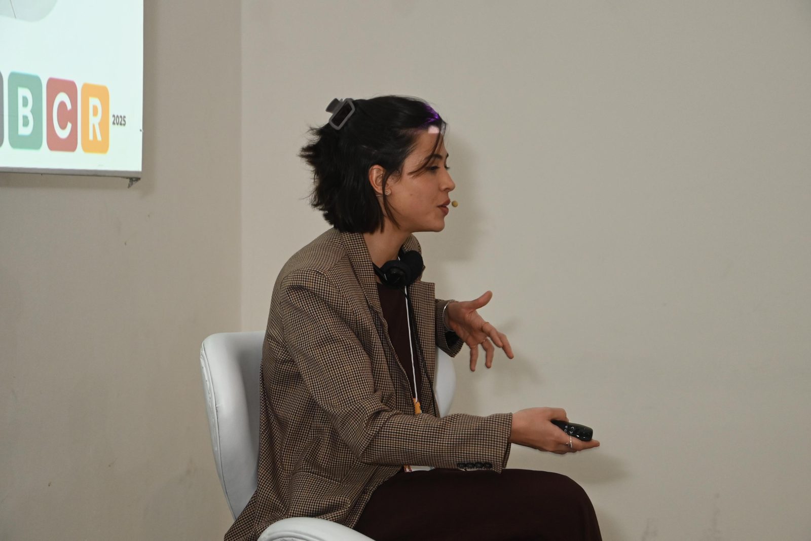 Integrante da equipe fala sentada num palco durante um painel, com a projeção de um evento ao fundo