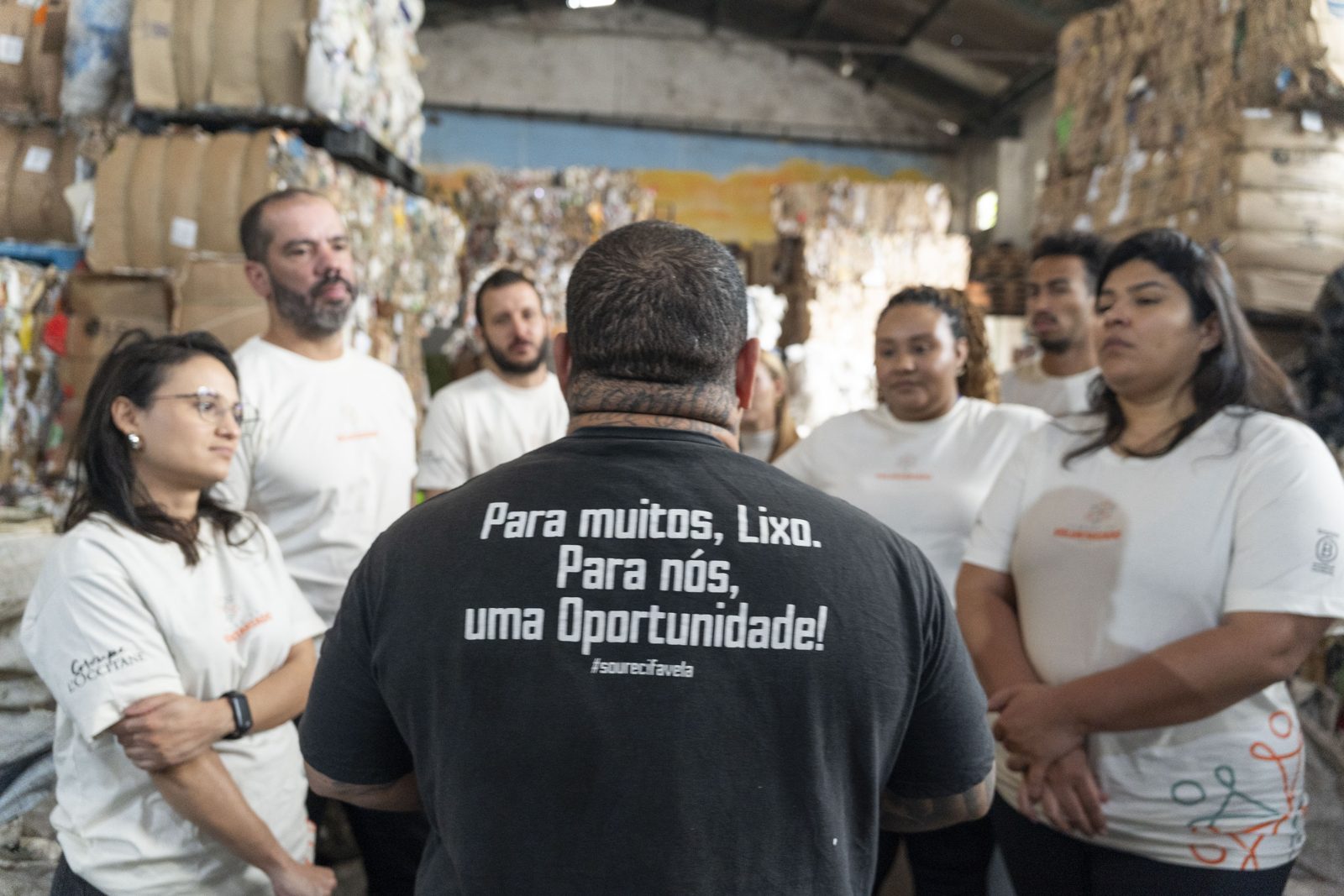 Liderança de uma cooperativa de reciclagem, de costas com a camiseta 'Para muitos, lixo. Para nós, uma oportunidade!', conversa com voluntários