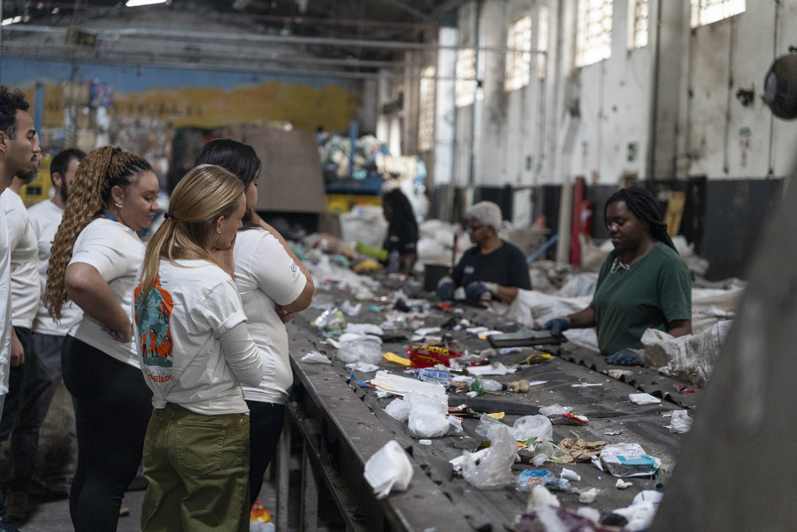 Voluntários observam catadoras separando materiais na esteira de triagem de uma cooperativa de reciclagem
