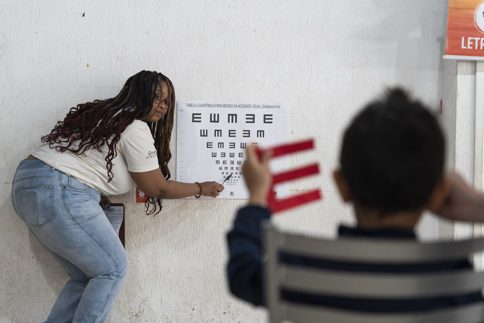 Voluntária aponta a tabela de acuidade visual durante um teste de visão com uma criança, que cobre um dos olhos