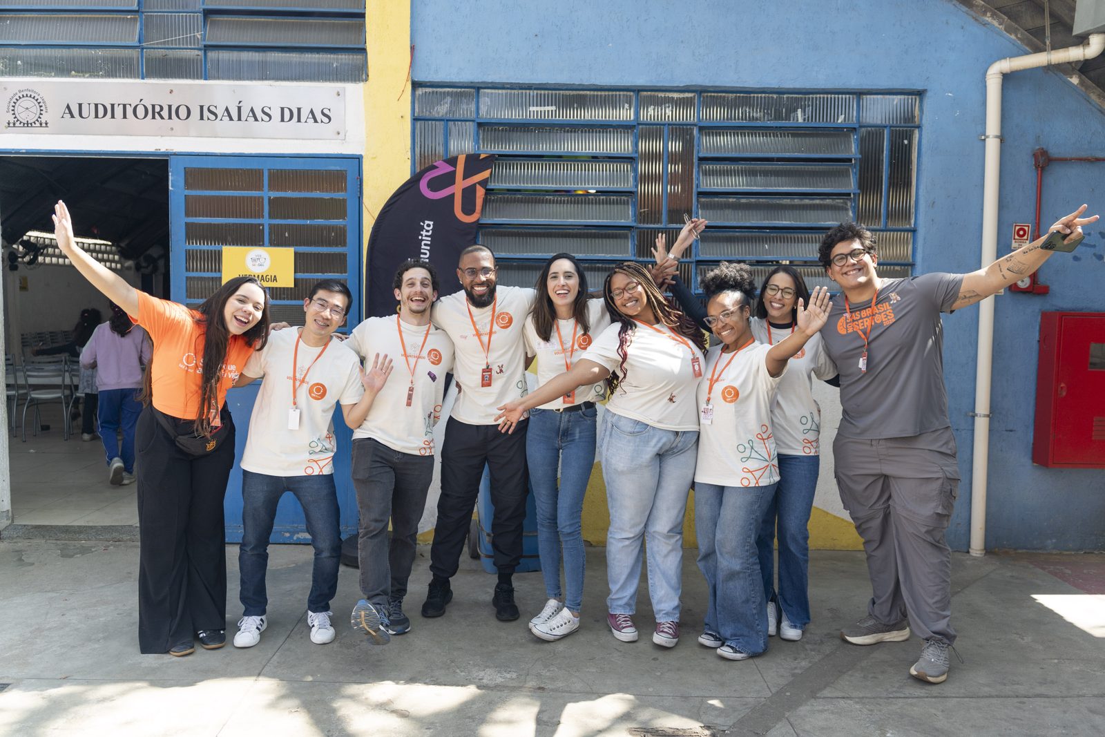 Grupo de voluntários sorridentes, de braços abertos, em frente ao Auditório Isaías Dias