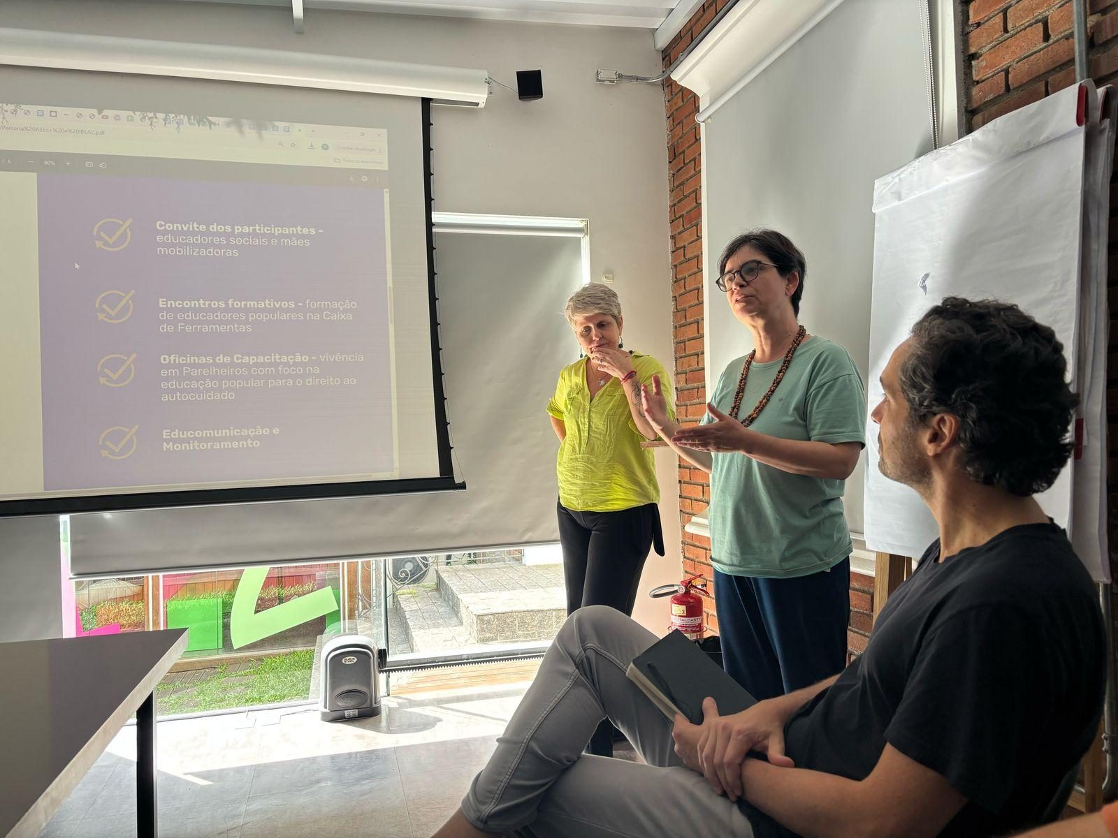 Facilitadora conduz uma conversa em frente à projeção, com participantes sentados ao redor
